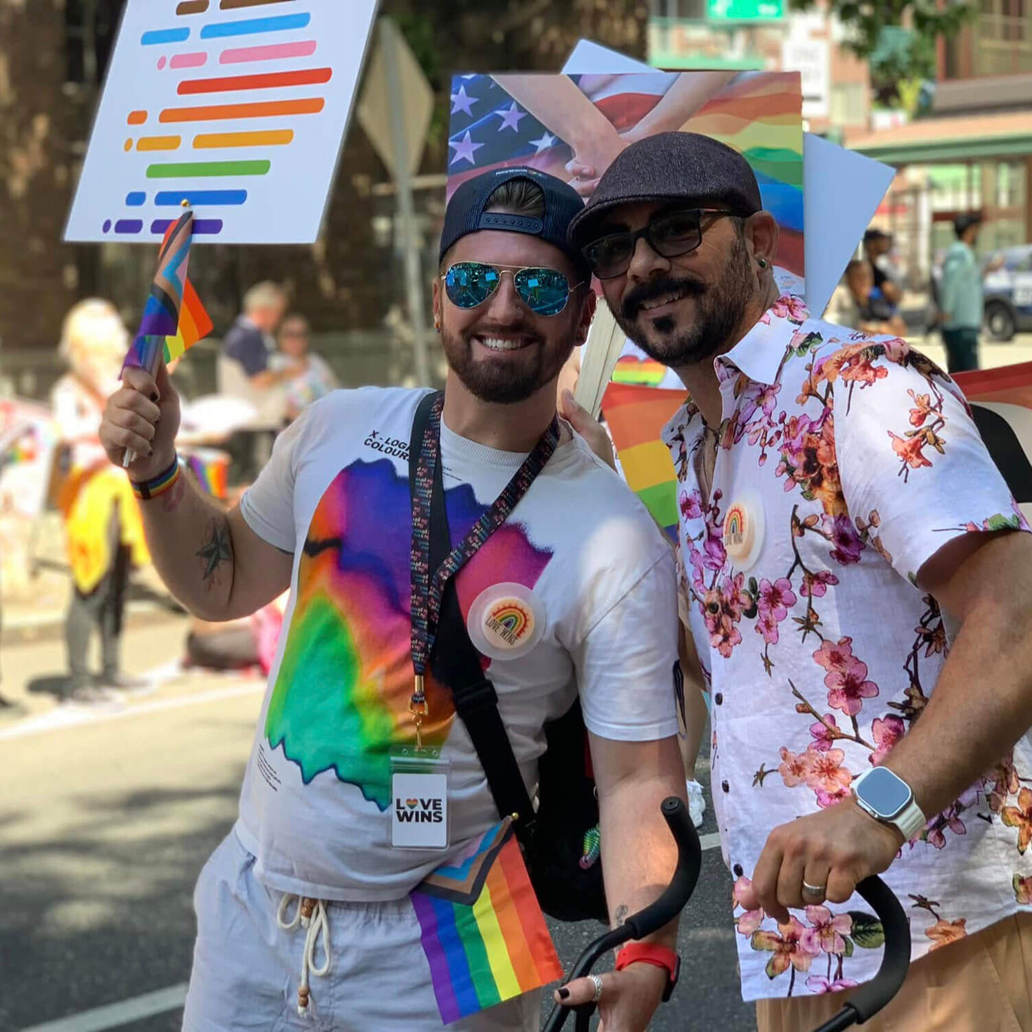 Two people smiling and posing together at a Pride parade, holding small rainbow flags
